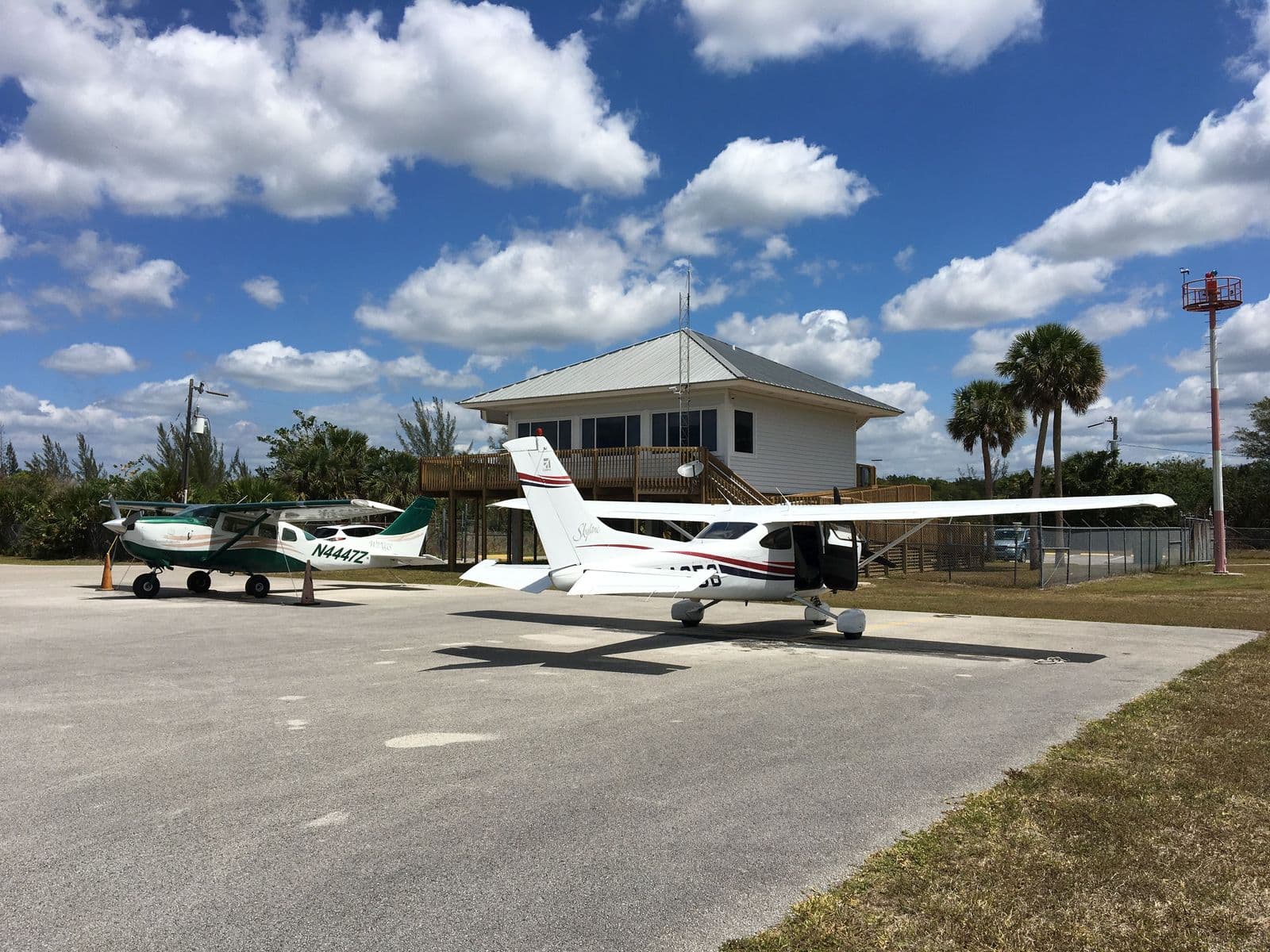Naše Cessna 182 Skylane s otevřenými dveřmi před budovou letiště Everglades Airpark, palmy a modrá obloha s mraky